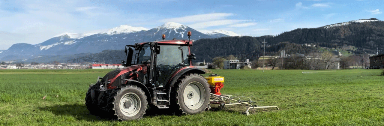 Traktor mit Nachsaatgerät arbeitet auf Grünland vor verschneiten Bergen Traktor mit Nachsaatgerät arbeitet auf Grünland vor verschneiten Bergen
